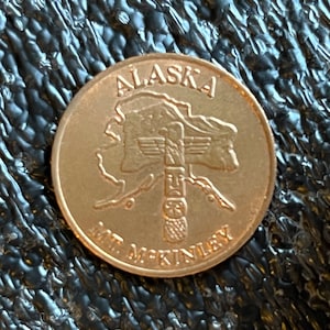 May include: A gold-colored coin with the words "Alaska Mt. McKinley" and an image of the state of Alaska with a totem pole in the center.