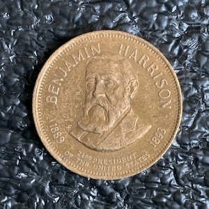 May include: A bronze coin featuring a portrait of Benjamin Harrison, the 23rd President of the United States. The coin is inscribed with "BENJAMIN HARRISON" at the top and "1889-1893" on either side. The text "23RD PRESIDENT OF THE UNITED STATES" is also visible.