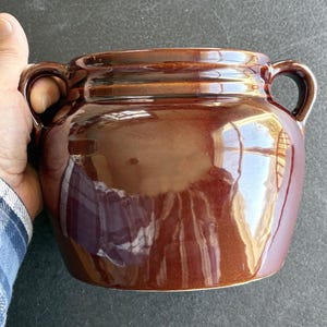 May include: A brown ceramic jar with two handles. The jar has a glossy finish and a rounded shape. The color is a rich, dark brown with lighter highlights. The jar is likely vintage or antique.