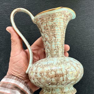 May include: A ceramic pitcher featuring a light mint green base color with a dark brown crackle glaze pattern throughout the body. The pitcher has a curved handle, a flared rim, and a gold-toned trim along the top edge and handle.