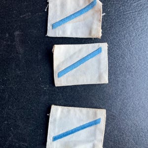 May include: Three vintage white fabric patches with a diagonal blue stripe. The patches have frayed edges and appear to be aged. The blue stripe is a solid color and runs across the center of each patch.