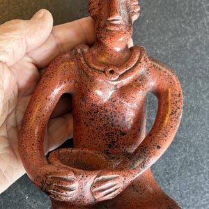 May include: A terracotta-colored figurine of a seated person with arms crossed. The figure has a stylized face and is adorned with a necklace. The surface has a speckled texture with black accents. The figurine is held in a hand.