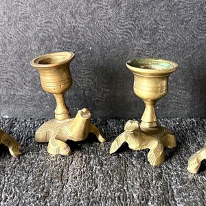 May include: A set of four brass candle holders, each featuring a turtle-shaped base with a textured shell and a central pedestal cup. The metal has a warm, aged gold tone with visible patina and green oxidation inside the candle cups.