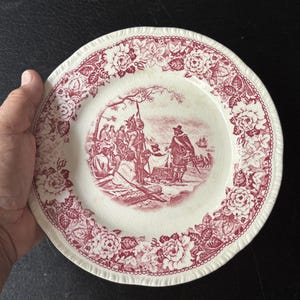 May include: A white ceramic plate with a red floral border and a central scene depicting figures in a historical setting. The plate has a scalloped edge. The plate is held by a hand.