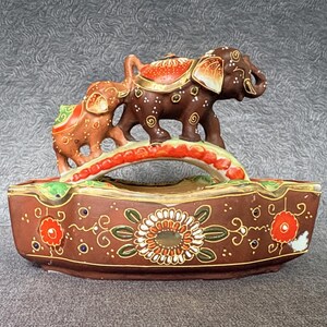 May include: A brown ceramic bowl featuring a decorative arched handle topped with two sculpted elephants. The bowl is adorned with hand-painted floral motifs in red, white, and green, accented with gold trim and small white dots on a dark brown base.