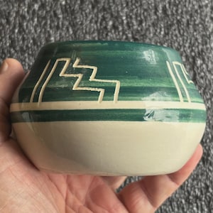 May include: A small, handcrafted ceramic bowl with a rounded base and a wide opening. The bowl features a cream-colored lower section and a green upper section with white geometric designs. The bowl is held in a hand.