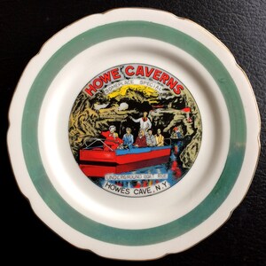 May include: A white ceramic plate with a green and gold rim. The center of the plate features a colorful illustration of a boat ride through a cave with the text "Howe Caverns Someplace Special Underground Boat Ride Howes Cave, N.Y."