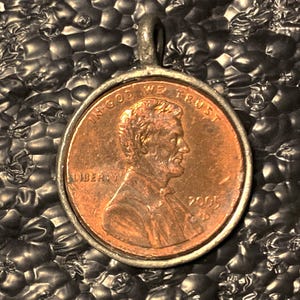 May include: A copper penny pendant featuring Abraham Lincoln's profile, dated 2005, set in a silver-colored metal frame. The coin displays the inscriptions "IN GOD WE TRUST" and "LIBERTY". The pendant has a small loop for a chain.