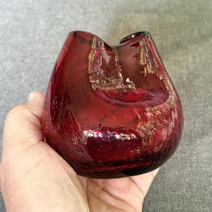 May include: A small, deep red glass vase featuring a distinct crackle texture throughout its surface. The vessel has a rounded, bulbous body with a wide, undulating rim that curves downward at the center, creating a unique, organic silhouette.