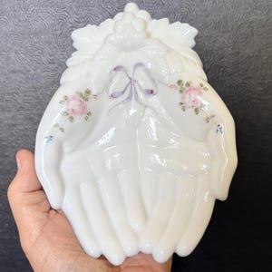 May include: A white milk glass trinket dish shaped like two cupped hands. The dish features a molded grape and leaf design at the top, a central purple ribbon detail, and delicate hand-painted pink rose floral accents on each side of the palms.