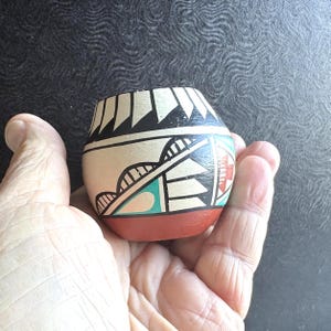 May include: A small, hand-painted ceramic pot with a rounded body and a flat base. The pot features geometric designs in black, white, red, and turquoise. The base is a solid reddish-brown color. The pot is held in a hand.