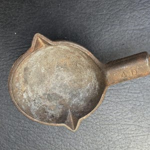 May include: A vintage, cast iron ladle with a round bowl and a handle. The ladle has a worn, rusty appearance, with the letters "A D" stamped on the handle. The bowl has a small spout and a pointed edge.