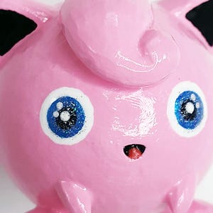 以下が含まれることがあります： 頭頂部にカールした髪の房があり、内側が黒い尖った耳、白いハイライトが入った大きな青い目、小さな口から赤い舌が見える、手塗りの丸いピンク色の架空の生き物のフィギュア。