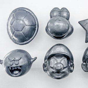 May include: A set of eight silver-colored character figurines from the Super Mario Bros. video game franchise. The figurines include Mario, Luigi, Bowser, Toad, a Goomba, a Koopa Troopa, a Bob-omb, and a star.