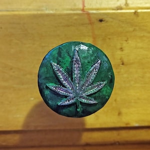May include: A circular drawer knob featuring a dark green, marbled finish with a raised, glittery, silver-toned cannabis leaf emblem centered on the front surface. The knob has a smooth, glossy texture and a rounded, slightly domed profile.