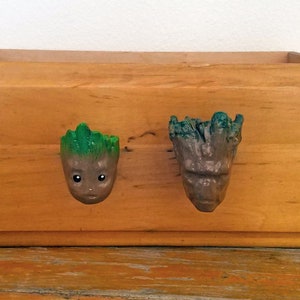 May include: Two wooden drawer pulls shaped like the character Groot from the Guardians of the Galaxy movie. The pulls are painted green, brown, and gray.
