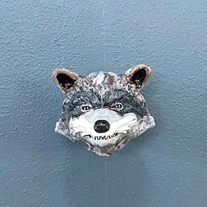 May include: A ceramic raccoon face drawer pull in grey and black with a white nose and mouth.