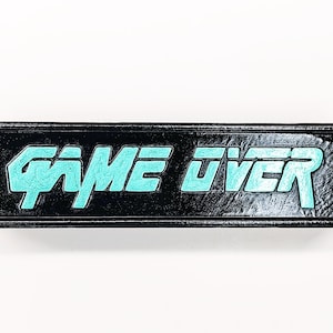 Op de afbeelding: Een rechthoekige zwarte plaquette met afgeschuinde randen met de tekst "GAME OVER" in een gestileerd, getextureerd lichtblauw lettertype. Het oppervlak van de plaquette heeft een licht ruwe, matte afwerking en de letters liggen verdiept.