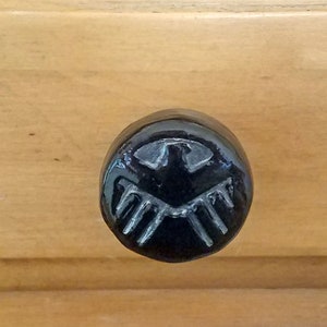 Peut inclure: Bouton de tiroir en céramique noire avec un motif de bouclier stylisé en relief.