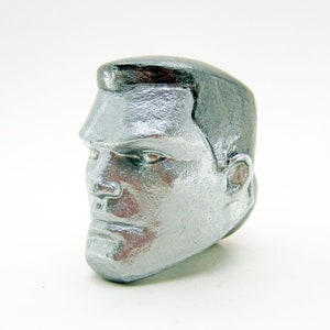 May include: A silver-coloured sculpture of a man's head. The sculpture has detailed facial features, including a prominent brow, defined nose, and a strong jawline. The hair is styled in a classic, swept-back manner. The sculpture is set against a white background.