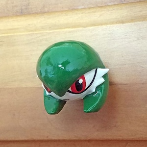 Könnte beinhalten: Ein grüner und weißer Schrankknauf mit einem Cartoon-Charakter-Design. Der Knauf hat eine glänzende Oberfläche und ein Gesicht mit roten Augen. Der Knauf ist an einer Holzschranktür befestigt.