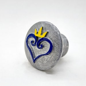 May include: A silver-colored metal knob with a blue heart and a yellow crown design.