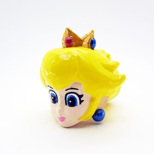 May include: A hand-painted figurine of Princess Peach from the Super Mario video game series. The figurine features a yellow head with blonde hair, blue eyes, a pink mouth, and a gold crown with red and blue jewels. The earrings are blue.