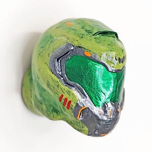 Può includere: Un casco verde con visiera verde metallizzata, accenti argentati e piccoli dettagli arancioni. Il casco presenta una finitura testurizzata e usurata con tre linee verticali rosse sul lato e una mentoniera di colore grigio scuro.