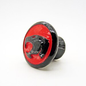 May include: A black and red knob with a black panther head design. The knob is round and has a small stem.