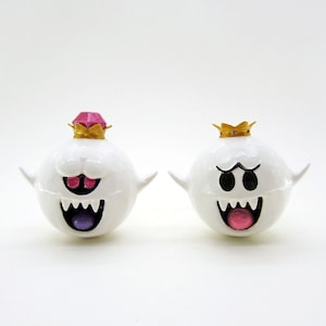 May include: Two white ghost figures with crowns and painted faces. One ghost has a pink crown and a purple tongue, the other has a gold crown and a pink tongue.