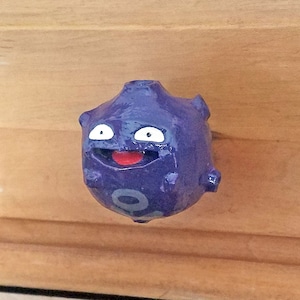 Könnte beinhalten: Ein violetter Schrankknauf in Form einer Cartoon-Figur mit einem lächelnden Gesicht, weißen Augen und einem roten Mund. Der Knauf ist an einer hellbraunen Holzschranktür befestigt. Der Knauf ist ein dekoratives Objekt.