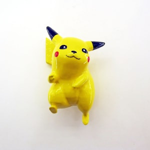 May include: A glossy yellow Pikachu figurine with black-tipped ears and red cheeks. The figure has a smiling expression and is a popular character from the Pokémon franchise. The figurine is made of a solid material.