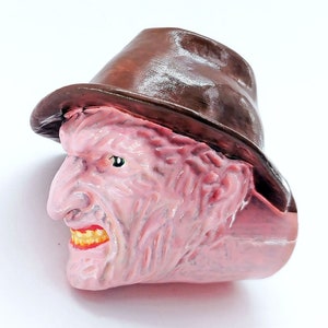 Peut inclure: Une figurine rose et brune d'un personnage de film d'horreur portant un fedora. Le personnage a une expression menaçante avec une grande bouche et des dents pointues.