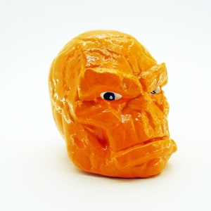 May include: Orange ceramic sculpture of a face with a furrowed brow and a menacing expression. The sculpture has a textured surface and blue eyes.