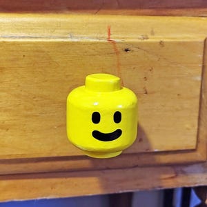 May include: A bright yellow cabinet knob shaped like a classic toy minifigure head. The knob features a smooth, cylindrical top stud and a printed black graphic of two oval eyes and a curved smiling mouth centered on the front surface.