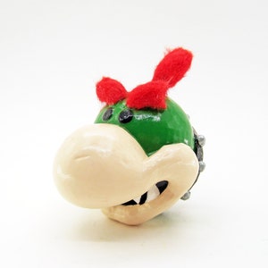 May include: A green and white ceramic figurine of a character with a red fuzzy hat and a toothy grin. The character has a spiky shell and large eyes.
