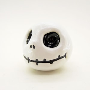 May include: White ceramic figurine of a character with a stitched mouth and one black eye. The character has a white face with black details.