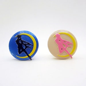 May include: Two round, 3D printed, decorative pins. One pin is blue with a black silhouette of a woman in a yellow crescent moon. The other pin is beige with a pink silhouette of a woman in a yellow crescent moon.