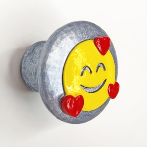 May include: A round cabinet knob with a textured silver finish featuring a yellow circular face with a smiling expression and three small, raised red heart shapes positioned around the perimeter of the yellow center piece.
