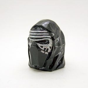 Puede incluir: Figura de casco de Kylo Ren negro con detalles plateados. El casco presenta un diseño detallado, que incluye una máscara y una capucha distintivas. La figura es un objeto de colección, probablemente de la franquicia Star Wars.