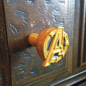 May include: Gold-coloured drawer pull with the Avengers logo. The logo is a stylised "A" with a circle around it.