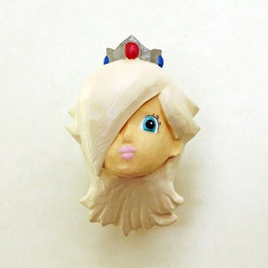 May include: A white figurine of a princess with a silver crown and blue and red gems. The princess has blonde hair, blue eyes, and pink lips.