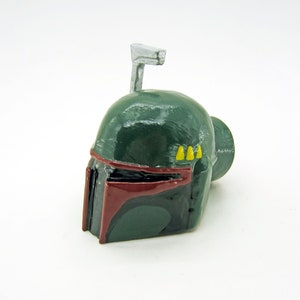 May include: A green and red plastic Boba Fett helmet with a silver antenna. The helmet has yellow details on the side.