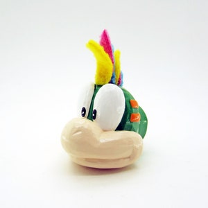 Peut inclure: Une figurine verte et blanche d'un personnage avec une crête de plumes colorées. Le personnage a de grands yeux blancs et une bouche grande ouverte.