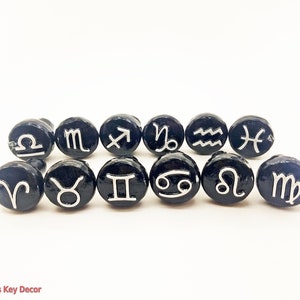 May include: Twelve black round ceramic tiles with silver zodiac symbols. The symbols are: Aries, Taurus, Gemini, Cancer, Leo, Virgo, Libra, Scorpio, Sagittarius, Capricorn, Aquarius, and Pisces.
