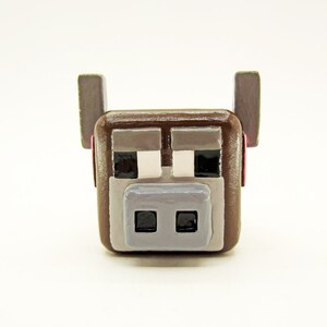 May include: A brown, square-shaped Minecraft-style cow figurine. The cow has gray and white details for the eyes and muzzle, with black square pupils. The ears are gray, and the overall design is blocky and pixelated.