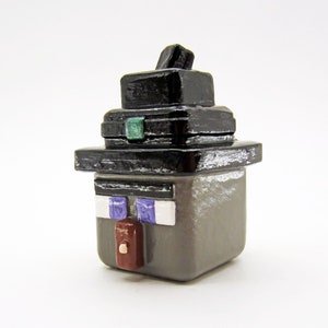 May include: A grey and black Minecraft figure with a black hat and a green gem on the front. The figure has a white face with purple eyes and a brown nose.