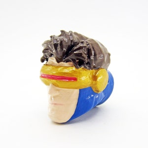 May include: A blue and gold plastic figure of a superhero with brown hair and a red visor. The figure is a head and shoulders portrait.