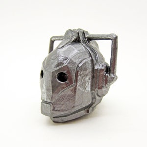 May include: A silver-colored, detailed, robot head figurine with a textured surface. The head has two black eyes and a raised, rectangular antenna.