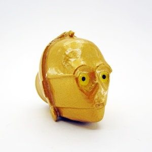 May include: A gold-coloured plastic figurine of C-3PO's head from Star Wars. The figurine has yellow eyes and a detailed face.